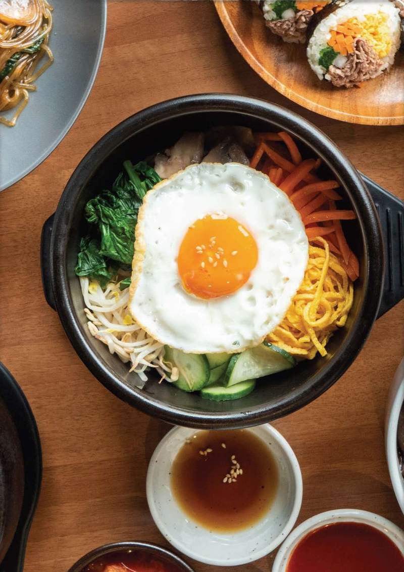 Dolsot bibimbap con uovo al tegamino, spinaci, carote e germogli, tra kimchi, salse e kimbab su un tavolo di legno