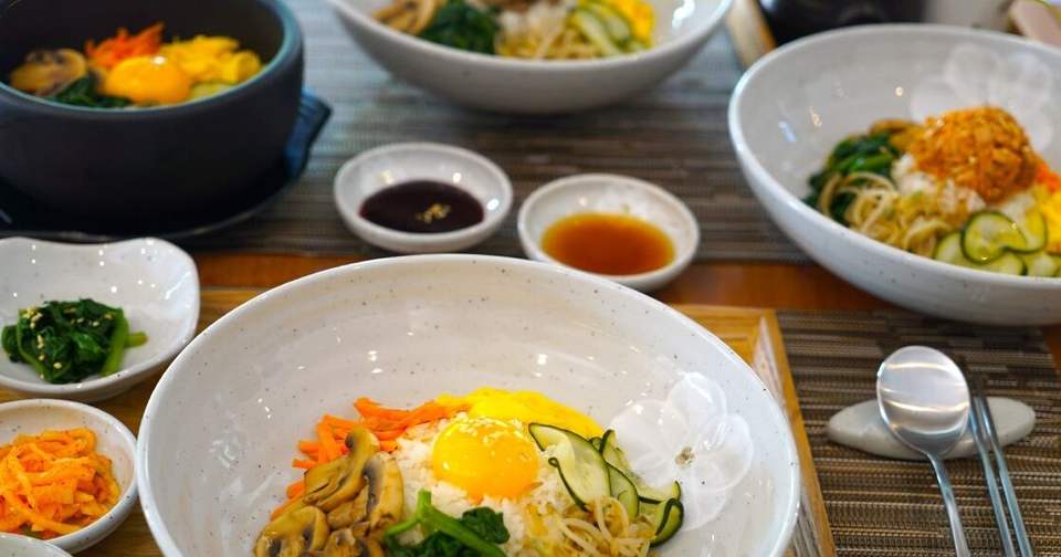 Bibimbap con uovo, funghi, spinaci e germogli in ciotola bianca, con banchan e dolsot sul tavolo del Dream
